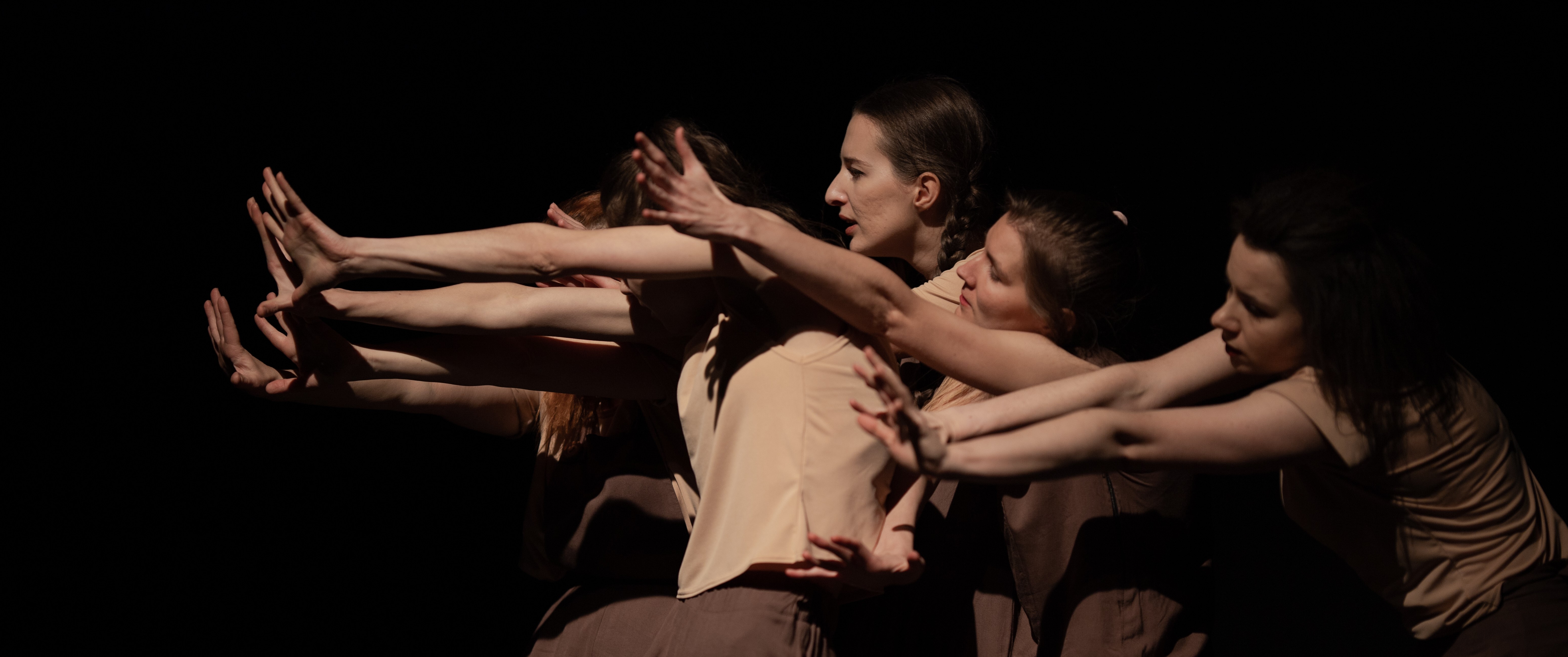 Photo shows group of young female dancers in beige and brown pushing left in a huddle on a darkened stage