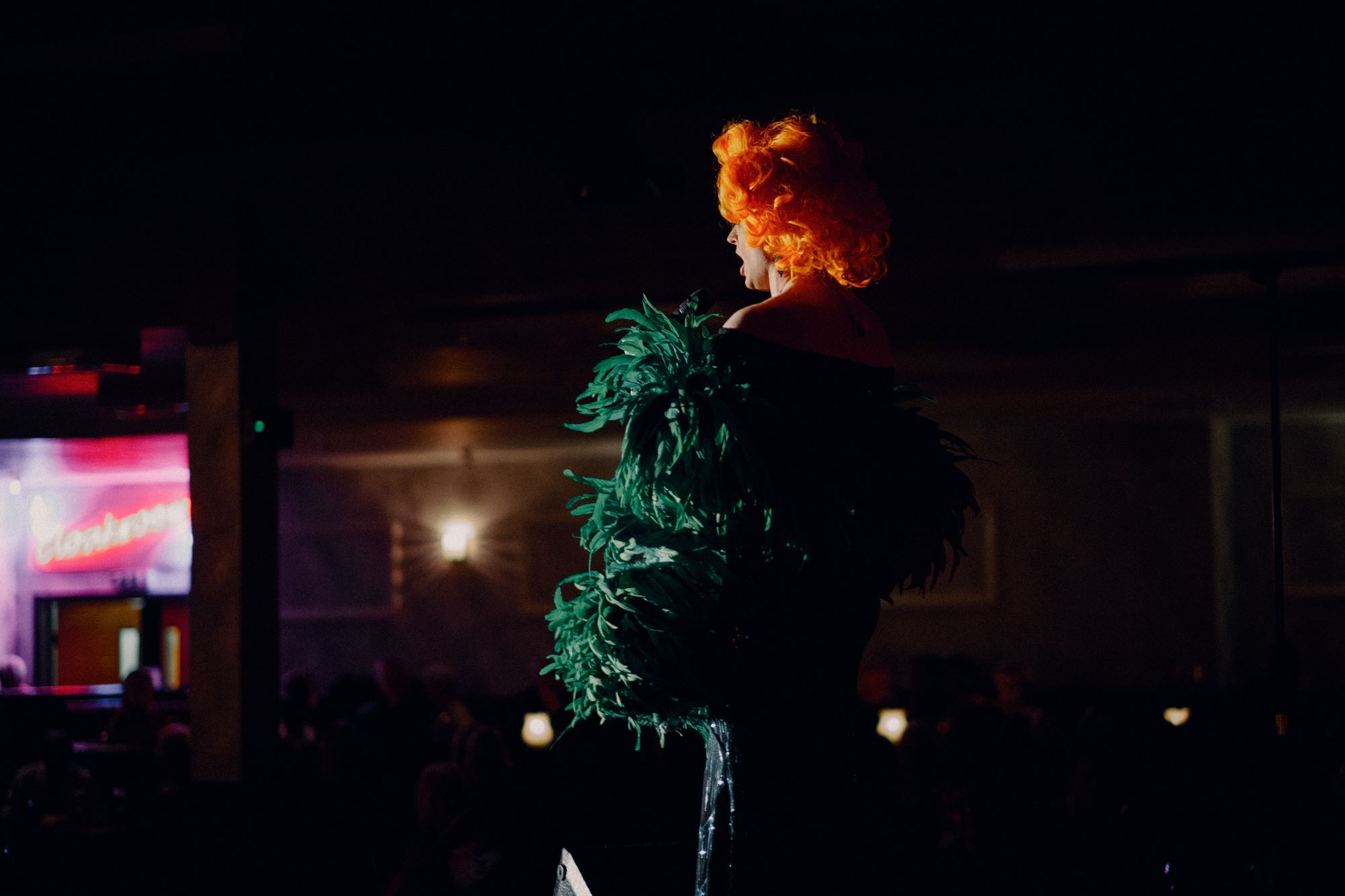 Photo shows drag artist in striking green feather dress and ginger wig performing on stage, the photo is taken from behind the artist in two thirds profile looking out towards the audience