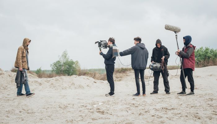 photo shows one hooded actor and young male camera crew filming on an empty sand dune
