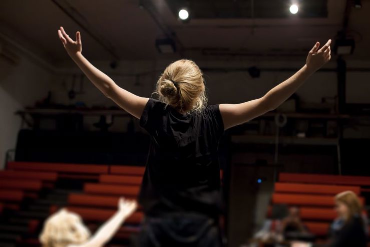 photo shows young woman from behind, she has her hands raised to the sky, she is facing the empty audience of a student theatre