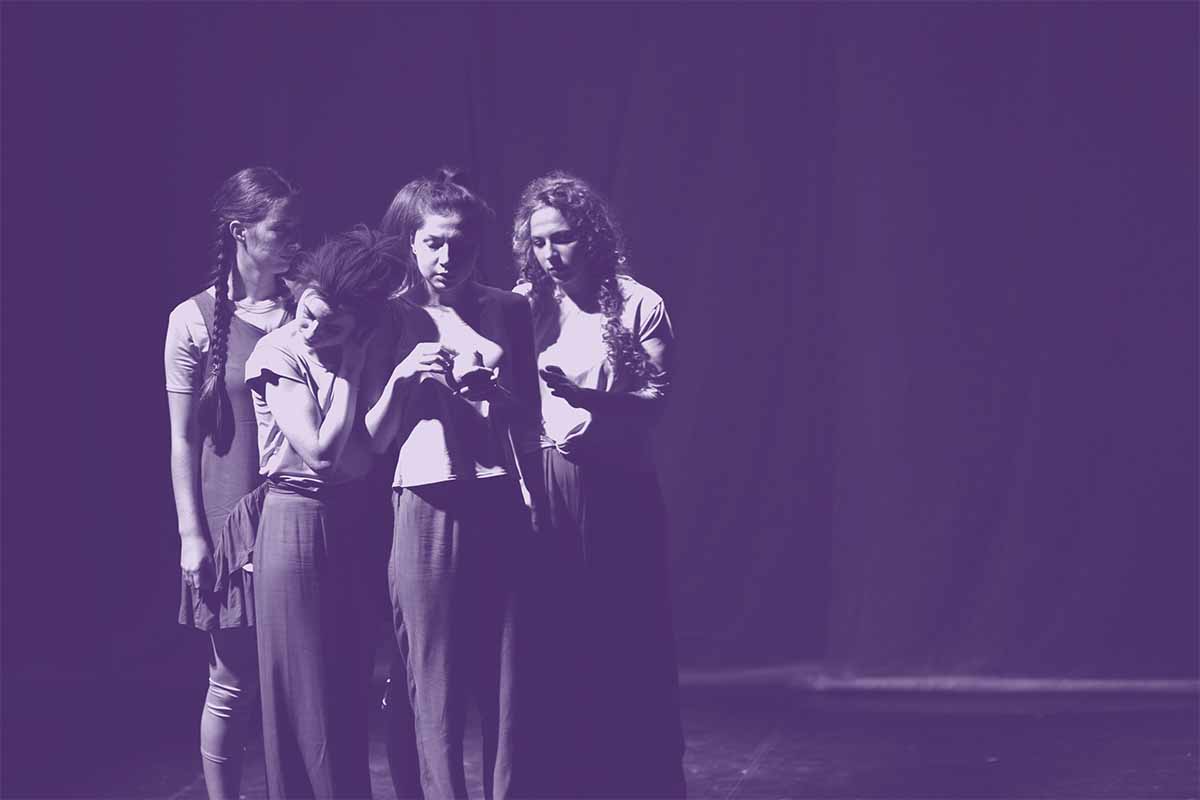 A group of performers huddle together on a bare stage