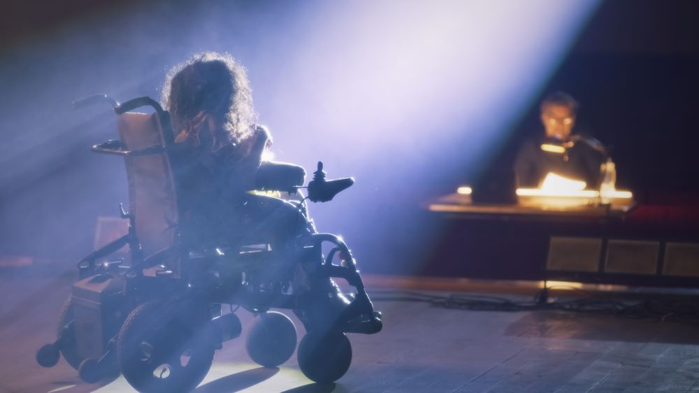 Photo shows an actor with a wheelchair on stage performing a monologue to onlooking director