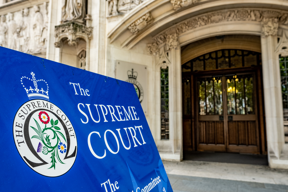 Photograph shows the entrance to the UK's Supreme Court building. The doors to the Neo-Gothic building are in the background and in the foreground is a blue placard that reads 'UK Supreme court'