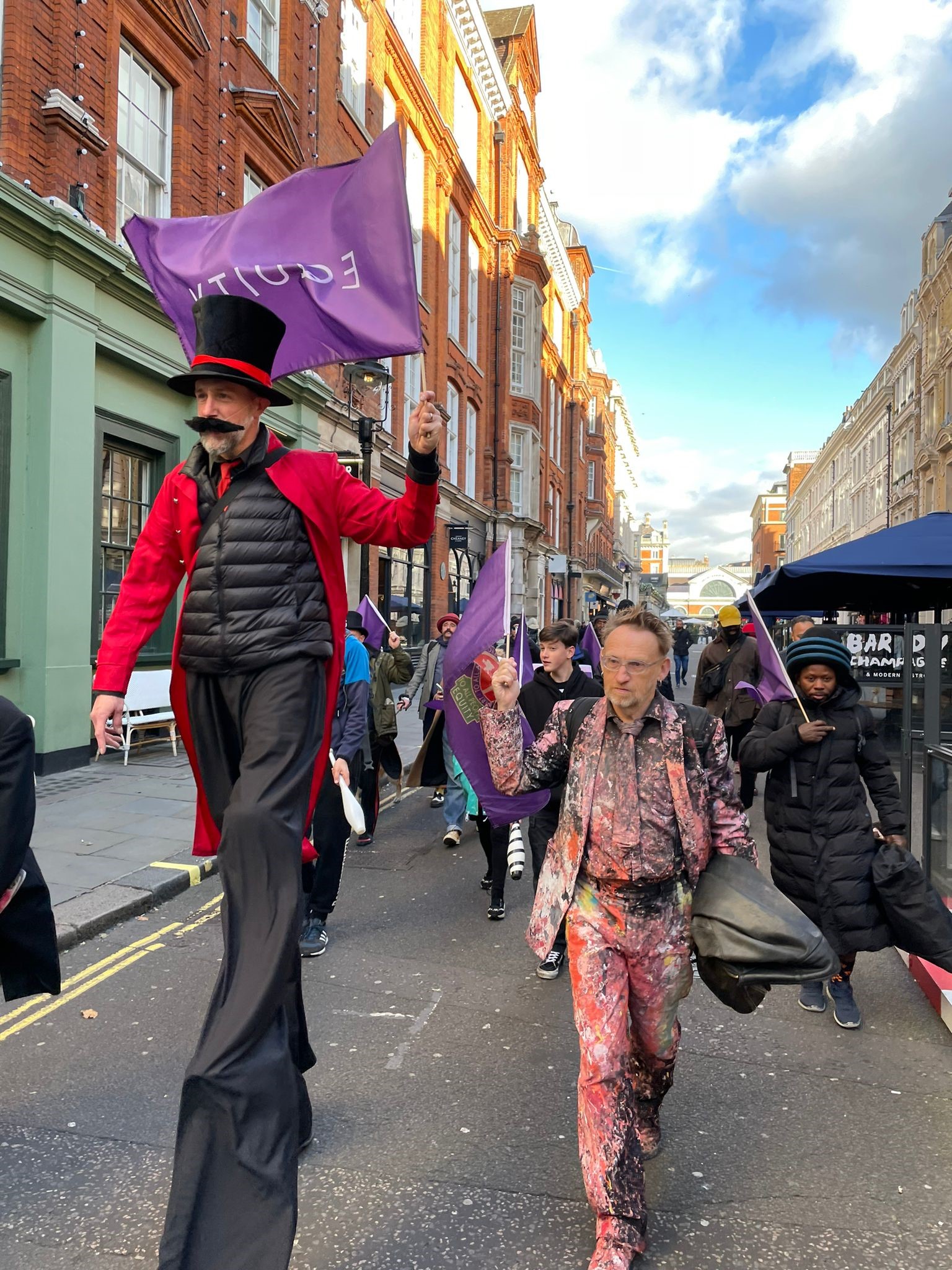 photo shows Equity member in ring master costume on slits walking through the streets of covent garden on slits followed by the street performers network waving equity flags