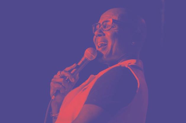 Photo shows black man with glasses doing a comedy set, the photo has a blue overlay
