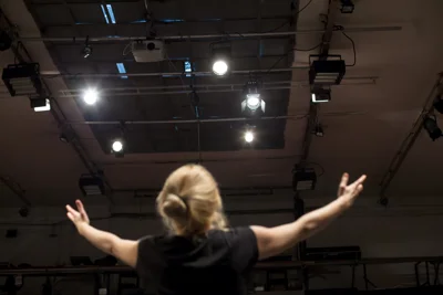 A singer with blond hair and arms outstretched, with back to the camera, sings under spotlights. 