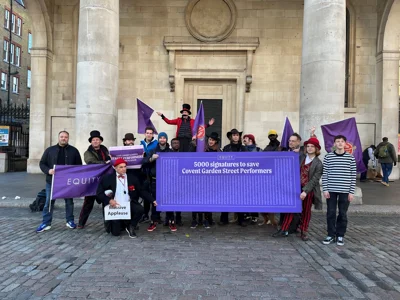 image shows group of around 20 Street Performers holding purple equity flags and a large petition of 5000 signatures printed on a large purple banner, outside St Paul's Church Covent Garden