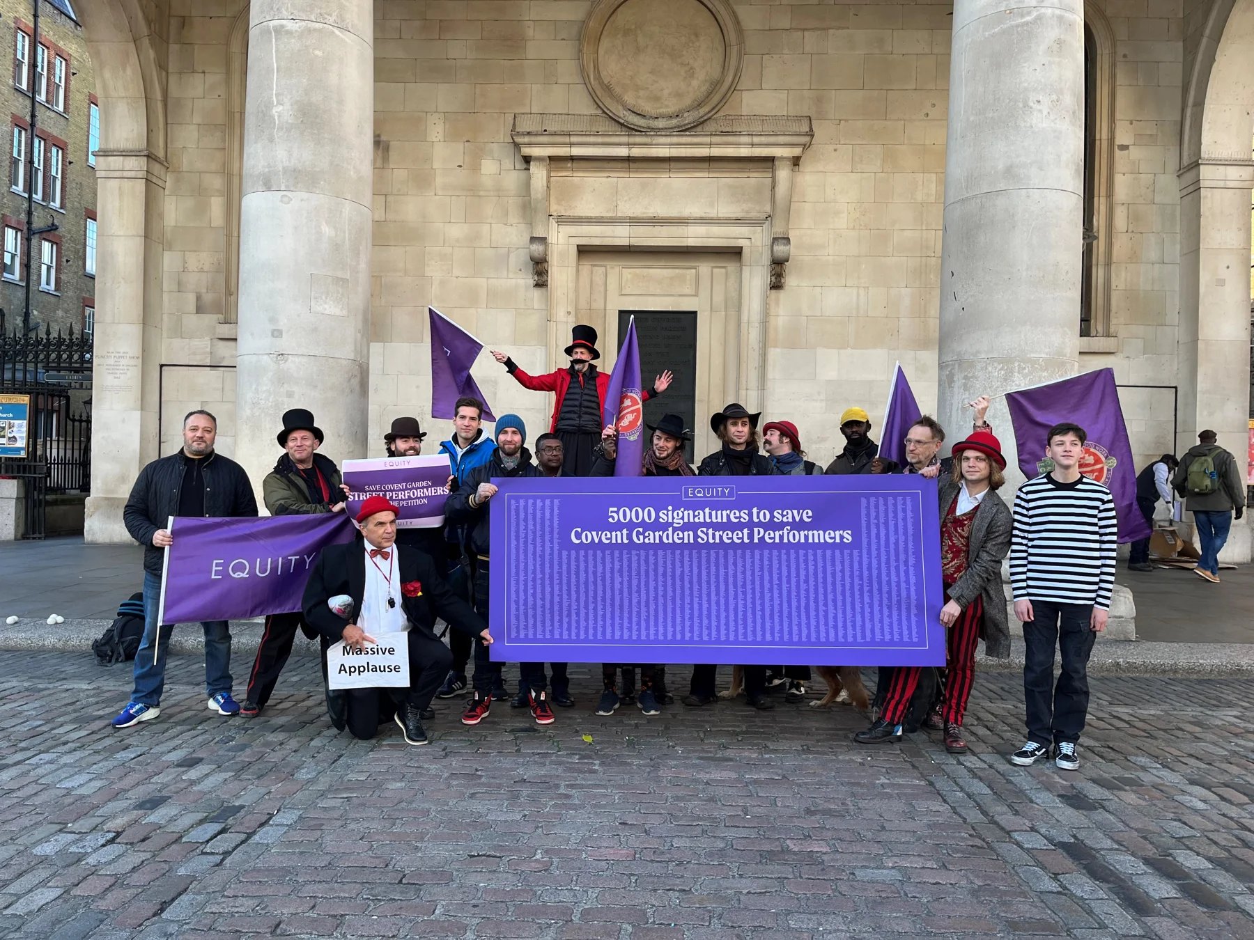 image shows group of around 20 Street Performers holding purple equity flags and a large petition of 5000 signatures printed on a large purple banner, outside St Paul's Church Covent Garden