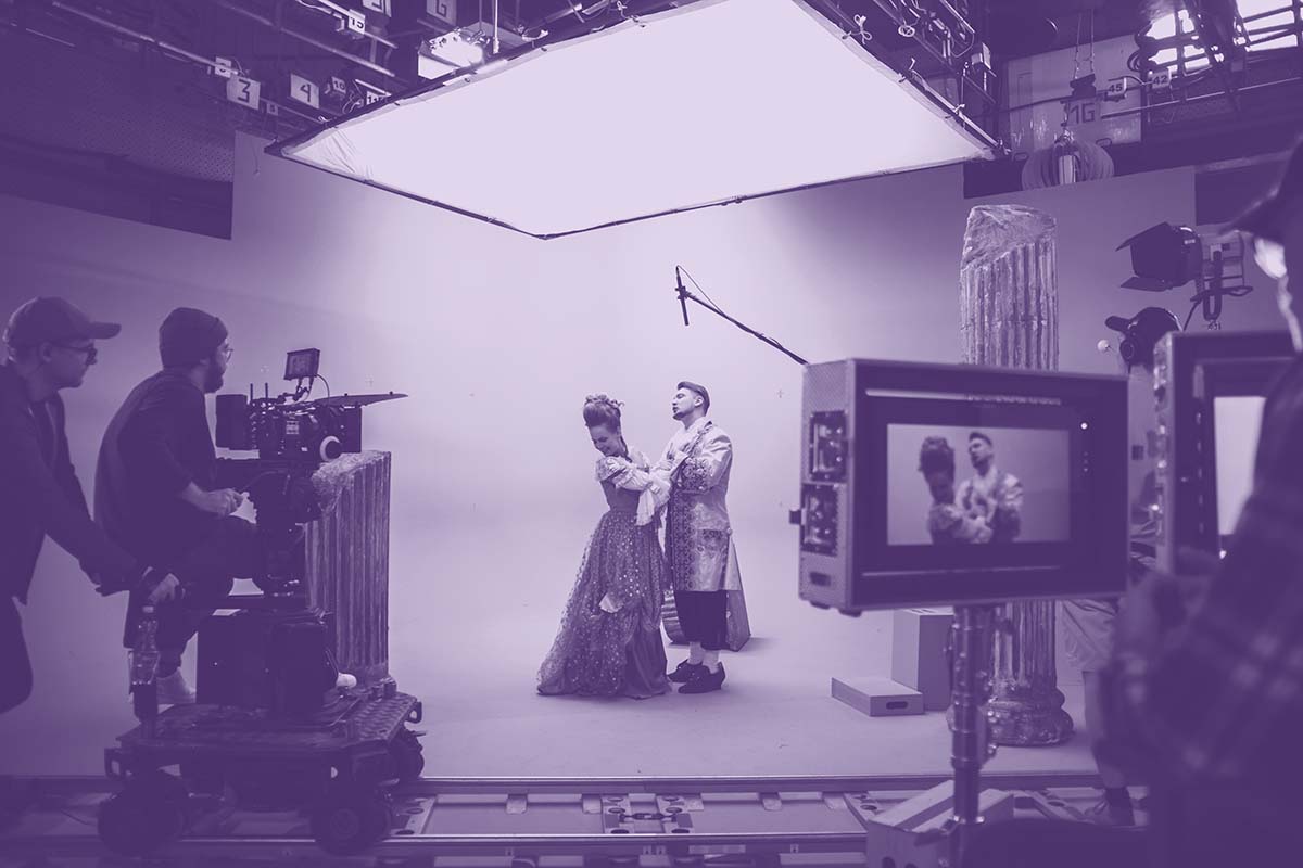 A film set, the focus is on a pair of actors in period costume in an embrace