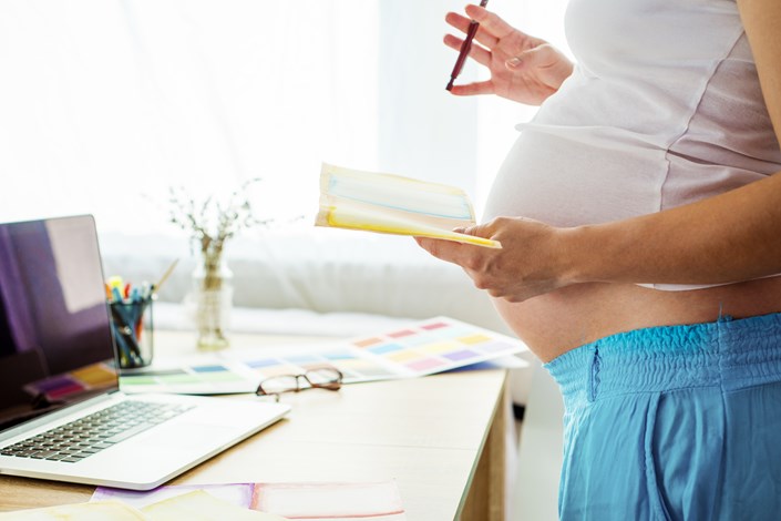 Pregnant woman in a home setting, stood at a desk with a laptop and notebook.